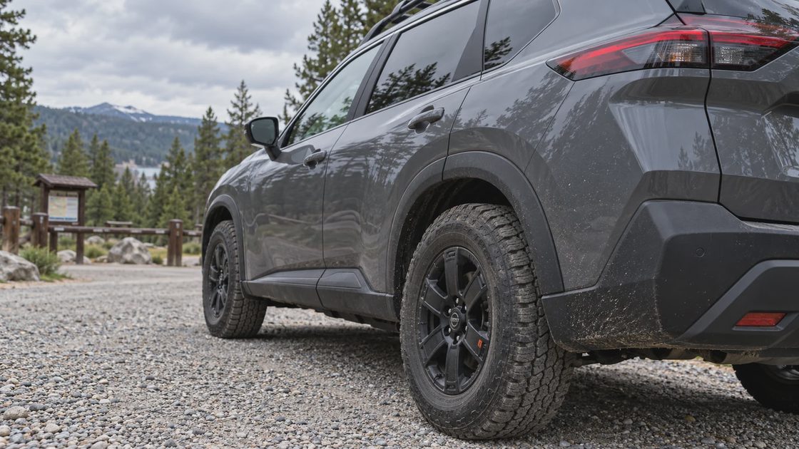 All-terrain tire and black wheel on a dark crossover at a gravel campground entrance.

